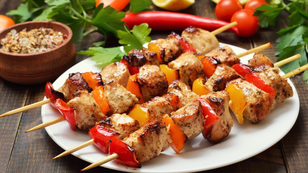 Grilled chicken and bell pepper skewers served on a white plate with a side of seasoning and fresh vegetables.
