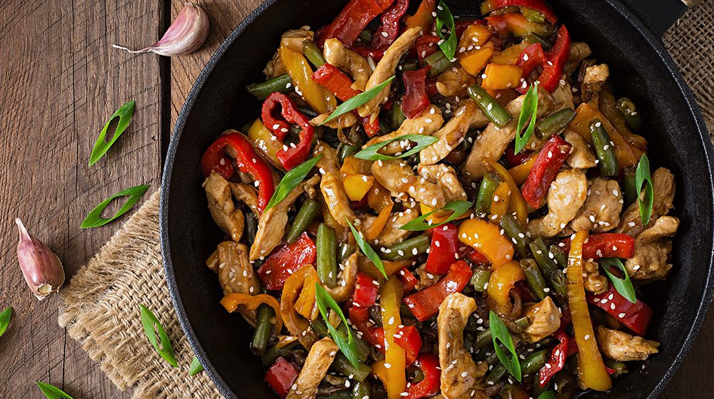 A vibrant stir-fry dish in a skillet with colorful bell peppers, green beans, chicken, and topped with sesame seeds and green onions.